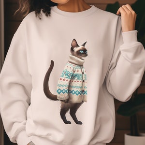 Può includere: Felpa bianca con un gatto siamese che indossa un maglione fantasia. Il gatto ha gli occhi blu e una coda marrone scuro. Il maglione ha una base color crema con disegni geometrici blu e rossi. Il gatto è in piedi.