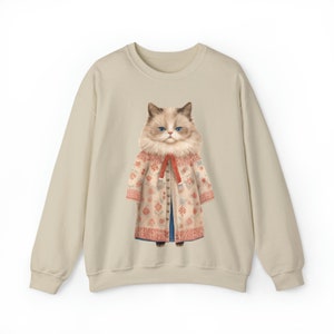 K&ouml;nnte beinhalten: Ein beiger Pullover mit einem grafischen Aufdruck eines Katers, der einen floral gemusterten Mantel und einen roten Schal tr&auml;gt.