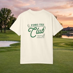 May include: Cream-colored t-shirt with the text "FORE-TEE Club" in green script and a golf club graphic. Below the club text is "AND STILL ON PAR ESTABLISHED 1986". The shirt is displayed on a golf course with a sunset in the background.