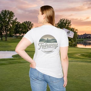 May include: White t-shirt with a circular graphic featuring a golfer, hills, and the text "Fairway Collection" in a vintage style. The shirt is worn by a person on a golf course with a flag in the background.