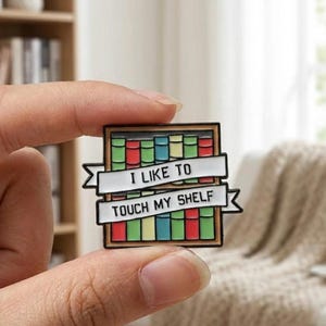 Può includere: Una spilla rettangolare in smalto raffigurante una libreria con libri colorati in rosso, verde e blu. Due striscioni bianchi si sovrappongono ai libri con il testo nero I LIKE TO TOUCH MY SHELF. La spilla ha un contorno in metallo scuro.