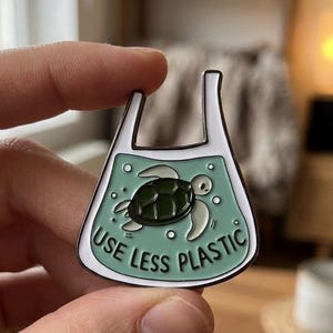 Peut inclure: Épingle en émail en forme de sac plastique, représentant une tortue de mer et le texte "USE LESS PLASTIC". L'épingle a un bord blanc, un fond vert clair et un motif de tortue vert foncé. Un accessoire pour les personnes éco-conscientes.