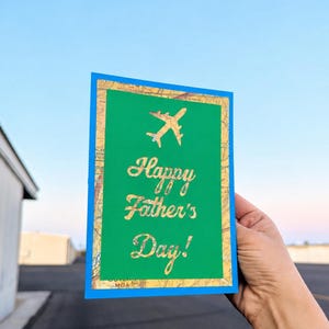 Peut inclure: Une carte de vœux pour la fête des pères avec un centre vert et un bord bleu. La carte présente un avion doré et les mots "Happy Father's Day!" en script doré. Le fond est un motif de carte.