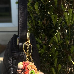 May include: A black quilted purse with a gold keychain featuring two detailed polymer clay charms. One charm depicts a colorful assortment of food items, while the other is a yellow circle with green and pink accents.