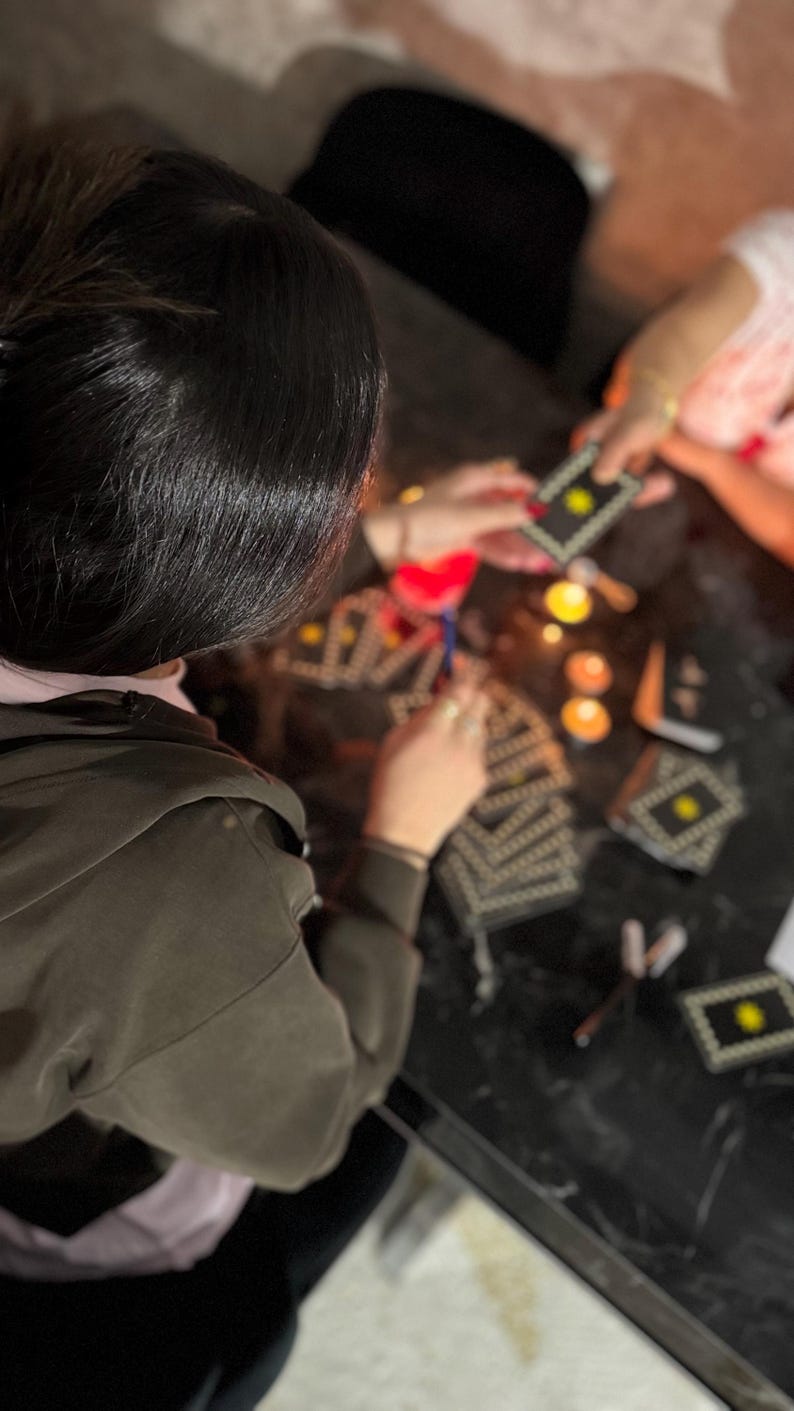 May include: A deck of rectangular tarot cards with black borders, white geometric patterns, and a central yellow star symbol. The cards are arranged on a dark surface alongside small lit candles, with some cards held in hands during a reading.