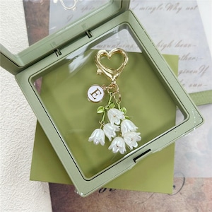 Peut inclure: Un porte-clés doré en forme de cœur avec une petite breloque circulaire blanche portant la lettre E en doré. Il comporte plusieurs breloques en forme de fleurs blanches en cloche avec des tiges et des feuilles vertes, suspendues à un fermoir mousqueton doré.