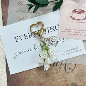 Peut inclure: Un porte-clés doré en forme de cœur avec un fermoir mousqueton, orné d'un bouquet de fleurs de muguet blanches aux tiges et feuilles vertes, accompagné d'une petite breloque ronde blanche portant la lettre G imprimée en doré au centre.