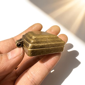 K&ouml;nnte beinhalten: Ein kleiner, rechteckiger Beh&auml;lter oder Anh&auml;nger aus Messing im Antik-Stil mit einer verwitterten, metallischen Oberfl&auml;che. Das Objekt verf&uuml;gt &uuml;ber einen Klappdeckel mit abgestufter, abgeschr&auml;gter Oberseite und eine kleine &Ouml;se an der Seite.