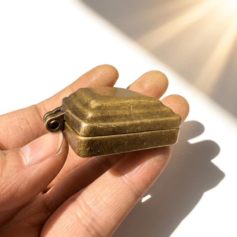 K&ouml;nnte beinhalten: Ein kleiner, rechteckiger Beh&auml;lter oder Anh&auml;nger aus Messing im Antik-Stil mit einer verwitterten, metallischen Oberfl&auml;che. Das Objekt verf&uuml;gt &uuml;ber einen Klappdeckel mit abgestufter, abgeschr&auml;gter Oberseite und eine kleine &Ouml;se an der Seite.