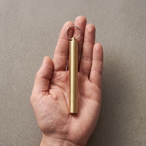 Puede incluir: Un pastillero cilíndrico de latón con un acabado mate liso y una base plana. La parte superior del recipiente cuenta con una pequeña anilla metálica integrada unida a una anilla plateada, lo que permite su uso como accesorio de llavero.