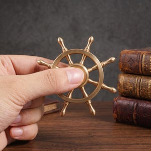 May include: A metallic gold-toned fidget spinner designed in the shape of a ship's steering wheel. The object features a central circular hub with eight radiating spokes that terminate in rounded tips, creating a nautical aesthetic.
