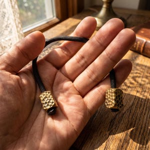 May include: A black cord featuring two cylindrical brass beads with a hammered, textured finish. The beads are positioned at each end of the cord, which is curved in a semi-circular shape. The metal has a warm, metallic gold-toned appearance.