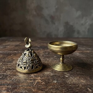 May include: A brass incense burner consisting of two separate pieces: a dome-shaped lid with intricate cut-out patterns and a small bird finial on top, and a matching pedestal bowl with a wide rim and a circular base.