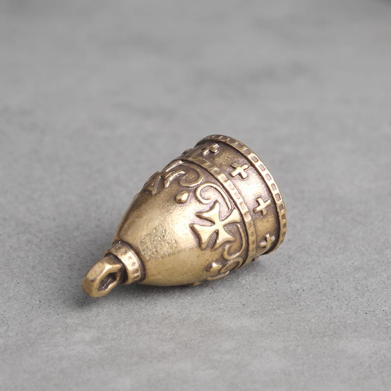 May include: A brass bell charm with a weathered finish, featuring embossed cross symbols and decorative scrollwork patterns around the exterior. The bell has a small loop at the top for attachment and a wide, open bottom edge with a textured border.