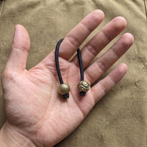 May include: A black paracord lanyard featuring two distinct brass beads. One bead has a textured, grid-like pattern, while the other bead displays a smooth, swirling, organic design. The cord is looped and secured with knots at the base of each bead.