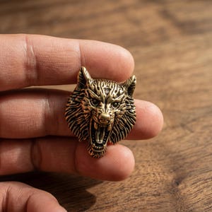 May include: A small, metallic, brass-coloured pin shaped like the head of a snarling wolf. The design features intricate, raised detailing that highlights the fur texture, pointed ears, and an open mouth with visible teeth, creating a high-contrast look.