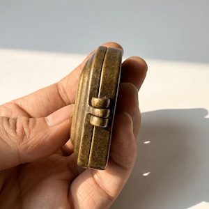 K&ouml;nnte beinhalten: Ein kleines, antik wirkendes Medaillon oder Etui aus Messing mit einem verwitterten, dunkelgoldenen Finish. Das Objekt verf&uuml;gt &uuml;ber einen markanten, au&szlig;en liegenden Scharniermechanismus an der Seite, der zwei abgerundete Metallh&auml;lften verbindet.