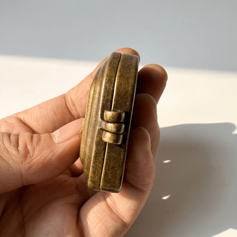 K&ouml;nnte beinhalten: Ein kleines, antik wirkendes Medaillon oder Etui aus Messing mit einem verwitterten, dunkelgoldenen Finish. Das Objekt verf&uuml;gt &uuml;ber einen markanten, au&szlig;en liegenden Scharniermechanismus an der Seite, der zwei abgerundete Metallh&auml;lften verbindet.