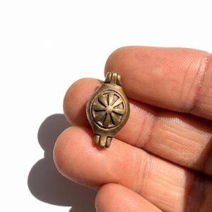 May include: A small, antique-style brass bead or charm featuring a central circular motif with eight radiating petals or spokes. The object has a weathered, metallic finish with a hinge-like structure at the top and bottom ends of the piece.