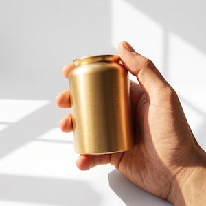 May include: A small, cylindrical container with a brushed gold finish and a smooth, rounded rim. The object features a metallic texture that reflects light, creating subtle highlights across its vertical surface and a curved top edge.