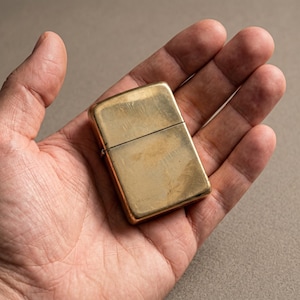 May include: A rectangular brass lighter with a polished, metallic gold finish and a hinged lid. The surface of the lighter shows subtle signs of wear and fine scratches, reflecting a warm, reflective tone across its smooth, flat exterior.