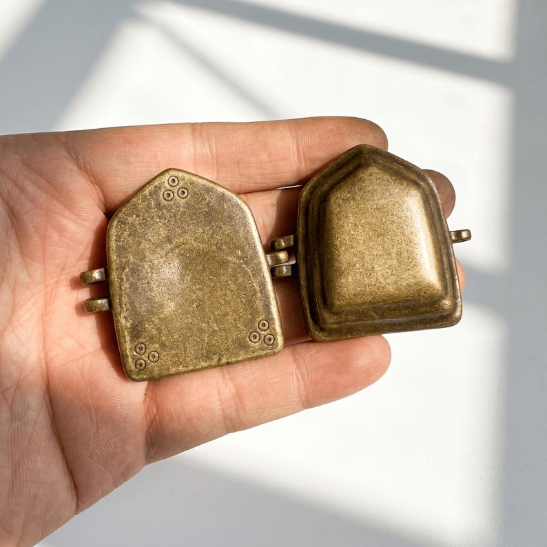 K&ouml;nnte beinhalten: Zwei messingfarbene Metallkomponenten in Form eines Spitzbogens. Ein Teil ist flach mit kleinen kreisf&ouml;rmigen Gravuren, w&auml;hrend das andere erhaben und kuppelf&ouml;rmig ist. Beide Teile verf&uuml;gen &uuml;ber kleine Metallschlaufen an den Seiten zur Verbindung.