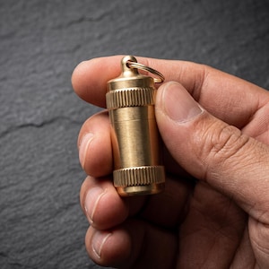 Può includere: Un contenitore cilindrico per pillole in ottone con tappo a vite caratterizzato da una texture zigrinata per una presa sicura. Il contenitore metallico presenta un piccolo occhiello sulla parte superiore collegato a un anello circolare.