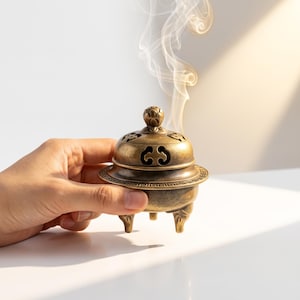 May include: A small, round, antique-style brass incense burner with three legs and a removable lid featuring intricate cut-out patterns and a lotus-shaped finial. Wisps of white smoke rise from the top of the metallic vessel, which has a weathered, golden-brown finish and a decorative border along the rim.