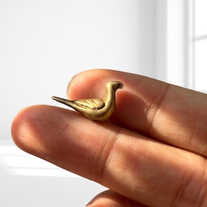 Könnte beinhalten: Eine kleine, goldfarbene Metallvogel-Figur mit einem glatten, abgerundeten Körper, einem spitzen Schwanz und einem gebogenen Hals. Die Oberfläche des Vogels weist feine, eingravierte Linien auf, die gefaltete Flügel andeuten.