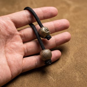 May include: A black paracord lanyard featuring two metallic beads. The top bead is shaped like a stylised owl head with detailed feather textures, while the bottom bead is spherical with a cross-hatched, textured surface finish in a brass tone.