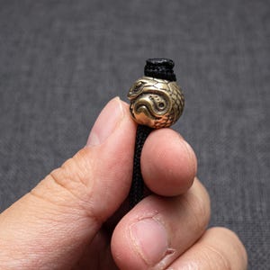 May include: A spherical brass bead featuring an embossed snake design with detailed scales and a coiled body, threaded onto a black braided cord. The metallic bead has a weathered, antique finish and a central hole for the cord to pass through.