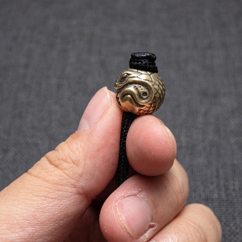 May include: A spherical brass bead featuring an embossed snake design with detailed scales and a coiled body, threaded onto a black braided cord. The metallic bead has a weathered, antique finish and a central hole for the cord to pass through.