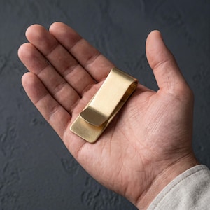 Op de afbeelding: Een rechthoekige geldclip gemaakt van geborsteld messing met een gladde, matte afwerking. Het metalen accessoire heeft een eenvoudig, gevouwen ontwerp met afgeronde hoeken en een egale, metallic gouden kleur over het gehele oppervlak.