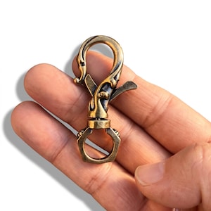 May include: A brass-toned metal lobster clasp featuring a curved, ornate hook design with a spring-loaded mechanism and a hexagonal swivel ring at the base. The hardware has a weathered, antique finish with dark recessed areas and polished highlights.