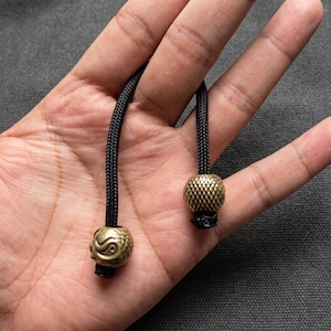 Könnte beinhalten: Zwei schwarze geflochtene Nylonschnüre, die jeweils mit einer metallischen Messingperle versehen sind. Eine Perle weist ein strukturiertes Schuppenmuster auf, während die andere ein detailliertes Spiraldesign zeigt. Diese Endstücke dienen als Zierde.