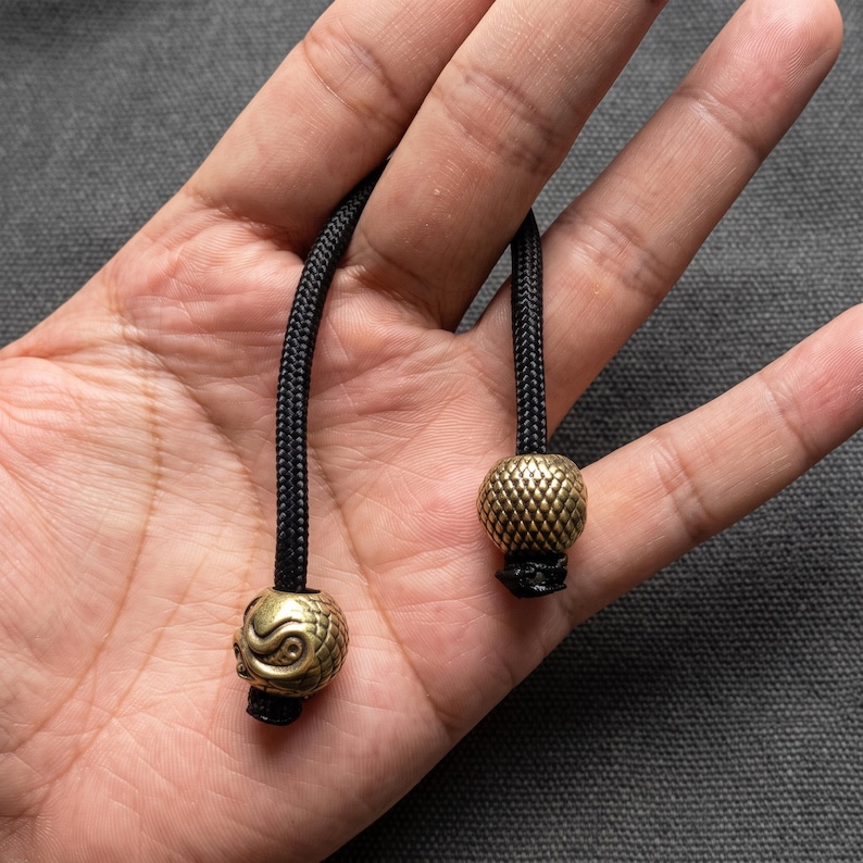 May include: Two black braided nylon cords, each finished with a metallic brass bead. One bead features a textured scale pattern, while the other displays a detailed spiral design. These decorative cord ends are designed for attachment to zippers.