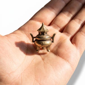 May include: A miniature brass incense burner featuring a rounded body, three short legs, two side handles, and a removable lid with a pointed top. The metallic surface has a weathered, antique finish with visible engraved detailing.
