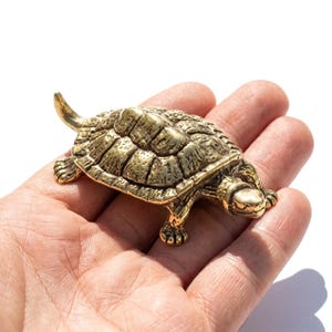 Op de afbeelding: Een klein, goudkleurig metalen schildpadbeeldje met een getextureerd, gesegmenteerd schild en vier gedetailleerde poten. De schildpad is afgebeeld in een wandelende positie met de kop naar voren gestrekt en een puntige staart aan de achterkant.