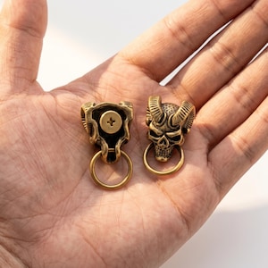 May include: Two brass-toned metal hardware pieces shaped like horned skulls. Each piece features a detailed skull with curved horns and a circular ring attached at the base. One piece shows a central screw head on the back side.