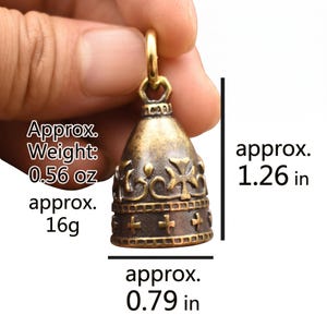 May include: A brass bell pendant featuring a cross and crown motif with a textured finish. The bell measures approximately 1.26 inches in height and 0.79 inches in width, with an approximate weight of 0.56 ounces, as indicated by the text.