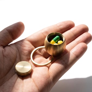 May include: A small, spherical brass pill container with a screw-top lid and an attached metal keyring. The container is open, revealing a green and yellow capsule and a round yellow pill inside, showcasing a brushed metallic finish.