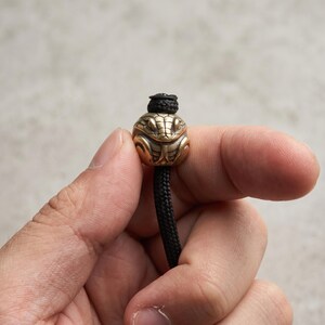 May include: A metallic, brass-toned bead shaped like a snake head with detailed scales and eyes, threaded onto a black braided cord. The bead features a rounded, compact design with a hole at the top and bottom for the cord to pass through securely.