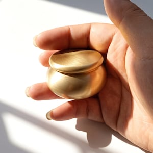 May include: A small, round, brushed gold-toned metal container with a smooth, domed lid. The object features a soft, matte metallic finish and a rounded, bulbous base, presenting a simple, compact, and circular design with a uniform golden color.