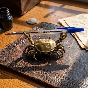 Op de afbeelding: Een messingkleurige metalen pennenhouder in de vorm van een krab, met opgeheven scharen die een doorzichtige BIC Cristal balpen met een blauwe dop en eindstuk ondersteunen. Het krabfiguur heeft een textuur op het schild en meerdere poten.
