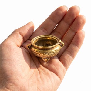 Könnte beinhalten: Ein kleines, rundes Räuchergefäß in metallischem Goldton mit zwei seitlichen Griffen und drei kurzen, dekorativen Füßen. Die Außenfläche der Schale ist mit komplizierten, geprägten Wirbelmustern und feinen Linien verziert.