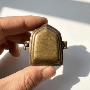 K&ouml;nnte beinhalten: Ein kleines, antik wirkendes Medaillon aus Messing mit einer spitz zulaufenden, bogenf&ouml;rmigen Oberseite und einer glatten, metallischen Oberfl&auml;che. Das Objekt verf&uuml;gt &uuml;ber ein Scharnierdesign an beiden Seiten und eine verwitterte Messingtextur.