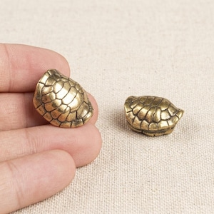 Puede incluir: Dos pequeños adornos metálicos de color dorado con forma de caparazón de tortuga, que presentan un patrón detallado de escamas hexagonales en relieve. Cada pieza tiene una forma redondeada y abovedada con un acabado pulido.
