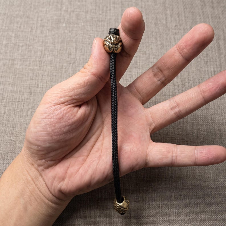 May include: A black paracord lanyard featuring two metallic bronze-toned beads. The top bead is shaped like a stylised animal head with intricate engraved details, while the bottom bead is spherical with a textured, repeating pattern.