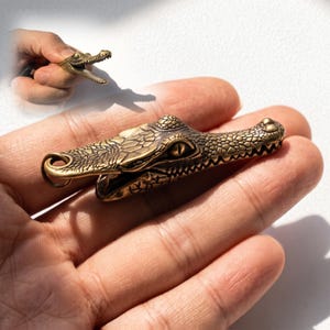 May include: A brass crocodile head charm featuring detailed scales, a textured surface, and a small metal ring at the back for attachment. The design captures the reptile's snout, eyes, and jawline with a metallic finish and dark patina accents.