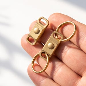 May include: Two gold-colored metal quick-release keychain connectors with a matte finish. Each piece features a central circular button mechanism and two rectangular slots at the ends, with one connector attached to a circular split ring.